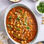 Chana Masala served in an oval light blue dish with raita, sliced onions, chopped cilantro and flatbreads.