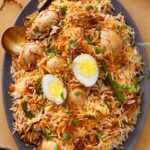 A platter of egg biryani garnished with boiled eggs, green chilies, fresh coriander, and crispy fried onions, served with a gold spoon on a tiled surface.