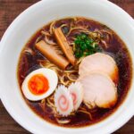 A beautifully plated bowl of shoyu ramen with chicken chashu, soft-boiled egg, menma, narutomaki, and scallions, served alongside rustic wooden chopsticks and spoon.