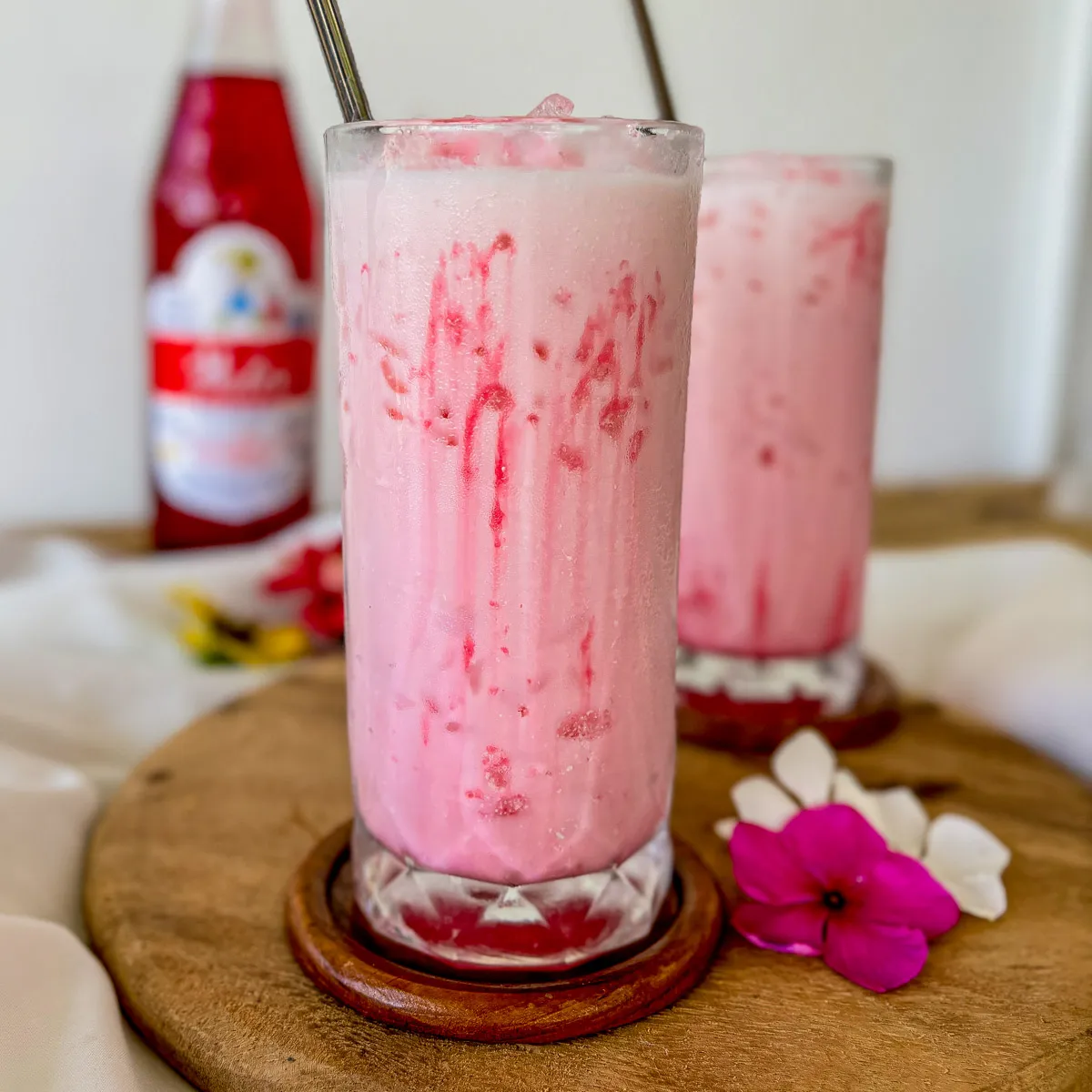 Nom yen, Thai pink milk, served in a tall glass with crushed ice and red syrup swirls, placed on a wooden coaster.