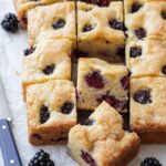 Cut squares of Blackberry Butter Cake on a piece of parchment, with a small blue knife and a few fresh blackberries scattered around.