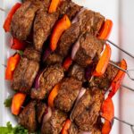 Broiled steak skewers on a platter with fresh parsley and creamy tahini sauce.