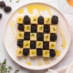 A plate of cheese cubes and blackberries arranged in a checkerboard pattern, garnished with herbs and drizzled with honey.