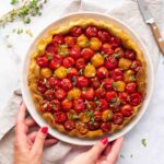 One person holds a plate containing a baked tomato tart, garnished with red and yellow cherry tomatoes and fresh herbs, placed on a marble surface, with a knife and napkin nearby.