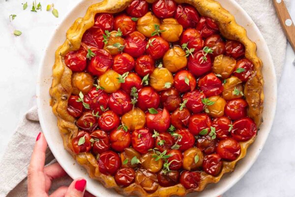 One person holds a plate containing a baked tomato tart, garnished with red and yellow cherry tomatoes and fresh herbs, placed on a marble surface, with a knife and napkin nearby.