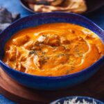 Side image of a blue bowl filled with creamy butter chicken curry sits on a table. Beside it are a plate with sliced naan bread, onions, and lemon wedges.