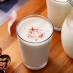 Creamy white sweet lassi topped with rose petals in a glass, placed on a blue & white tiled table.