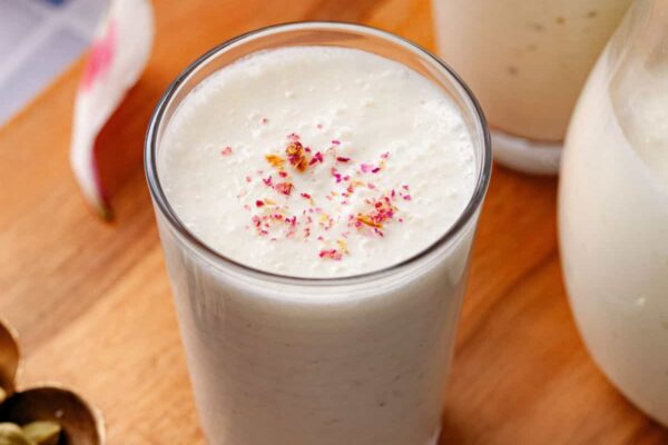 Creamy white sweet lassi topped with rose petals in a glass, placed on a blue & white tiled table.