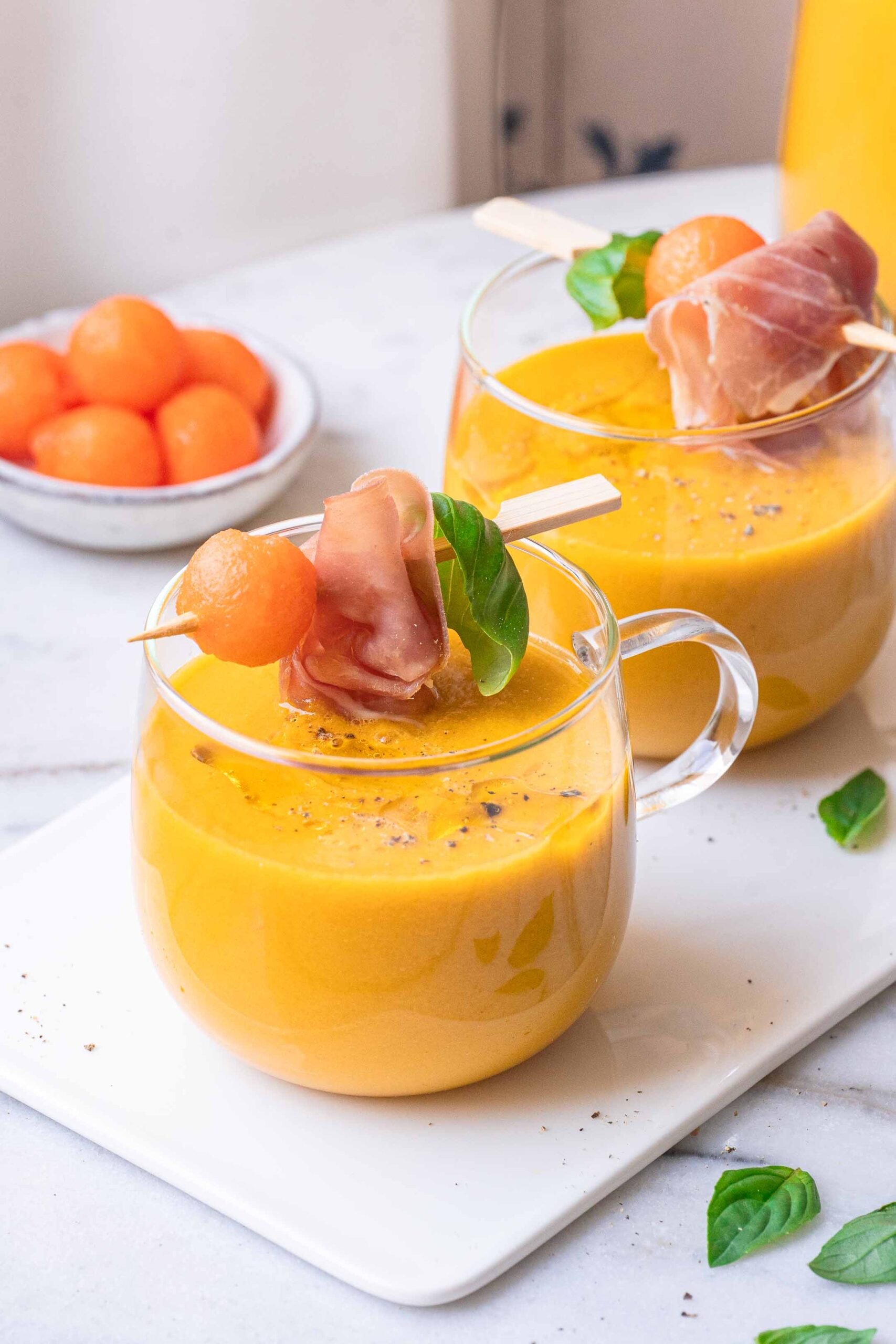 Cold Melon Soup with Basil and Mint 1 Two glasses of orange melon soup are garnished with prosciutto skewers, basil and melon balls. A bowl of melon balls is in the background.