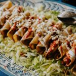 Crispy homemade chicken flautas served on a bed of shredded lettuce in a blue and white hand-painted dish, topped with red chile sauce, a drizzle of Mexican crema, and crumbled queso fresco – a delicious take on a classic chicken flautas recipe.