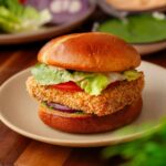 A crispy breaded paneer burger with lettuce, tomato, and sauce on a toasted bun, served on a plate with sliced vegetables and sauce in the background.