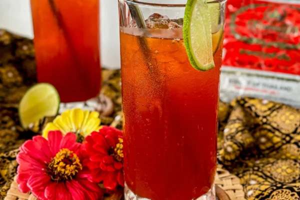 Cha manao, Thai lime iced tea served in a tall glass with ice and metal straw, surrounded by fresh flowers on a woven coaster.