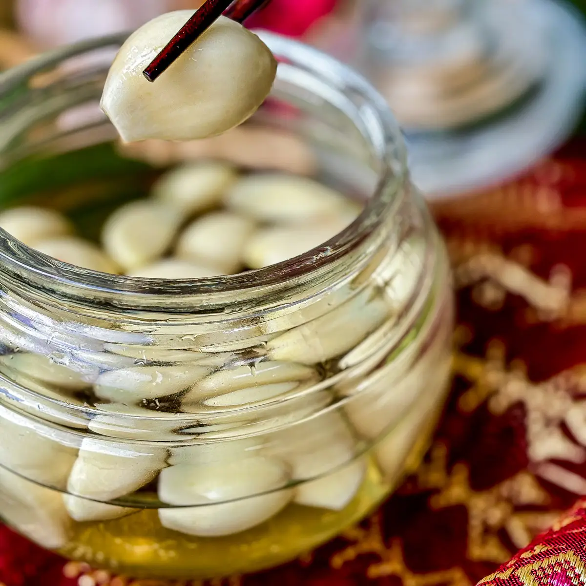 A jar of pickled garlic with clear brine and one clove being lifted by chopsticks.