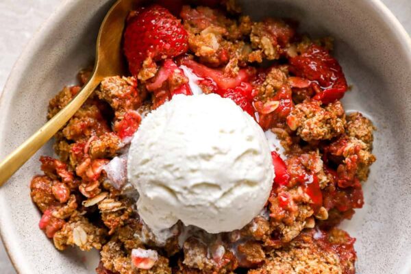 A bowl of strawberry crisp topped with vanilla ice cream with a spoon on the side.