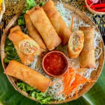 Thai egg rolls with spicy dipping sauce in a traditional bamboo basket, garnished with fresh lettuce, fried noodles, and shredded carrots.