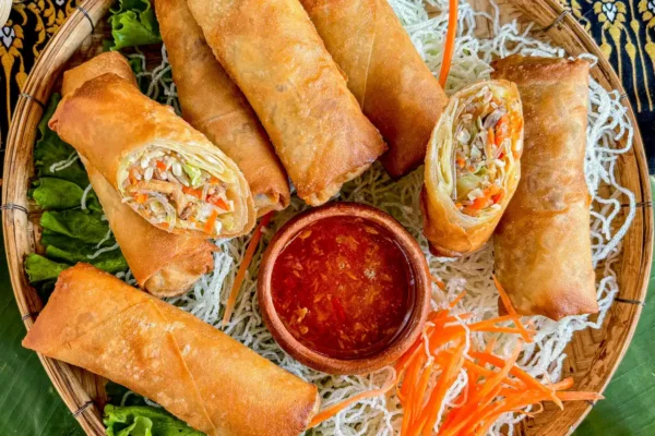 Thai egg rolls with spicy dipping sauce in a traditional bamboo basket, garnished with fresh lettuce, fried noodles, and shredded carrots.