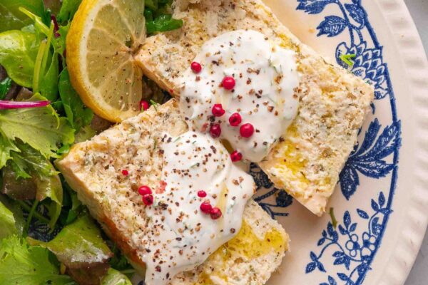Two slices of bread topped with cream and pink pepper, accompanied by mixed greens and a slice of lemon on a blue and white floral plate.