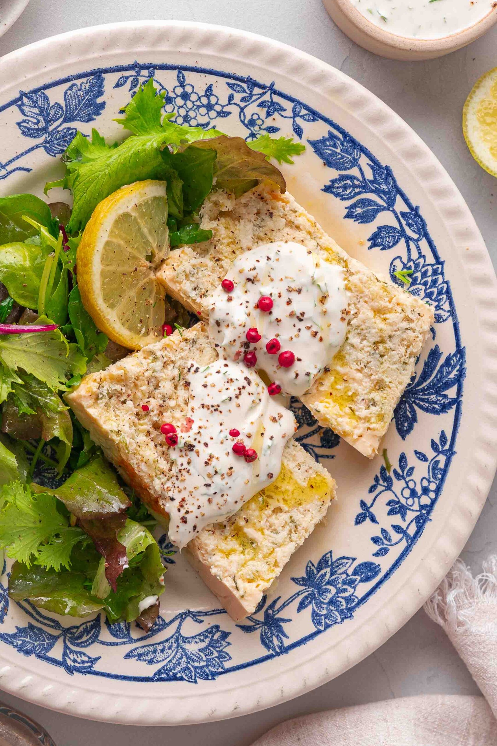 Two slices of bread topped with cream and pink pepper, accompanied by mixed greens and a slice of lemon on a blue and white floral plate.