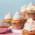 Sprinkling rainbow sprinkles over a batch of frosted funfetti cupcakes