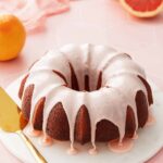 Pink glazed grapefruit bundt cake on a marble cake plate with a pink background and a cut pink grapefruit in the background.