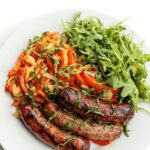 Grilled bratwursts with grilled peppers and onion on a plate with arugula and garnished with basil ribbons.