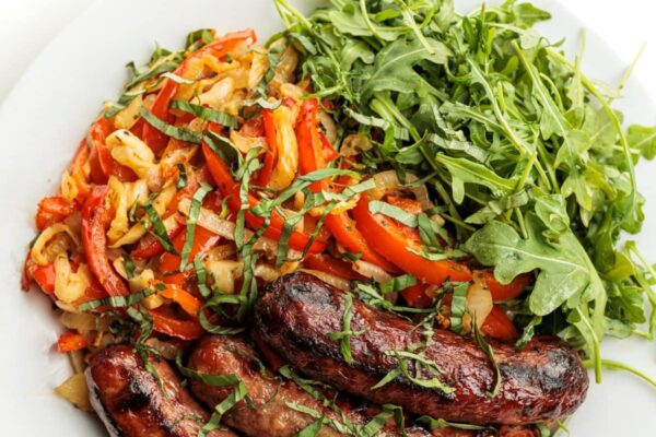 Grilled Bratwurst with Peppers and Onions Grilled bratwursts with grilled peppers and onion on a plate with arugula and garnished with basil ribbons.
