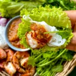 Close-up of grilled Thai shrimp lettuce wrap with peanut sauce and rice noodles held above a platter of fresh ingredients.