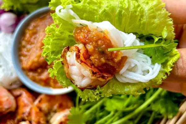 Close-up of grilled Thai shrimp lettuce wrap with peanut sauce and rice noodles held above a platter of fresh ingredients.