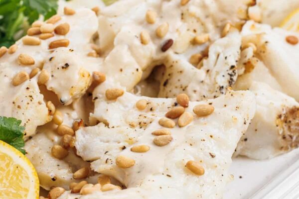 Close up view of grilled white fish with tahini lemon sauce and garnished with toasted pine nuts.