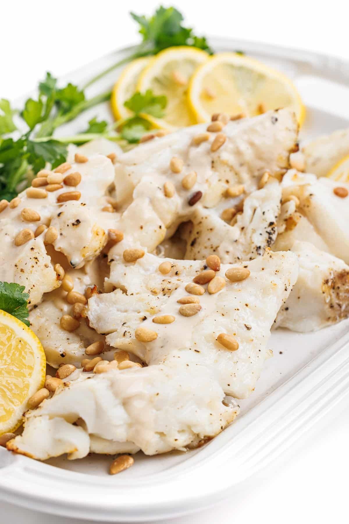 Close up view of grilled white fish with tahini lemon sauce and garnished with toasted pine nuts.