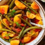 Greek green beans stew in a gray-brown mottled bowl garnished with dill and with a silver spoon ready to eat. The bowl stands on a dark wooden surface. In the background you can see a slice of baguette and a small bowl of Tzatziki.