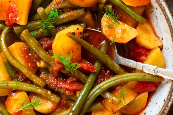 Greek green beans stew in a gray-brown mottled bowl garnished with dill and with a silver spoon ready to eat. The bowl stands on a dark wooden surface. In the background you can see a slice of baguette and a small bowl of Tzatziki.