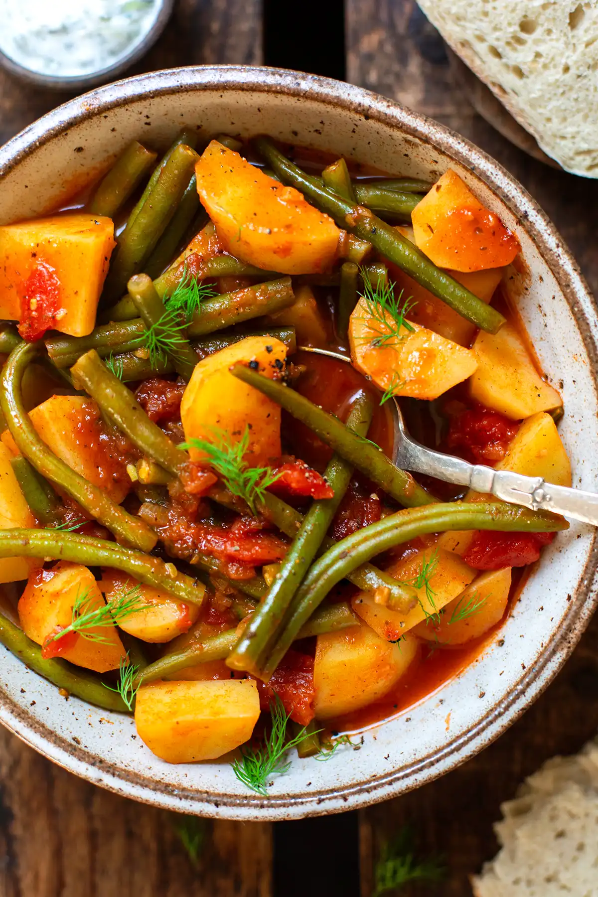 Greek green beans stew in a gray-brown mottled bowl garnished with dill and with a silver spoon ready to eat. The bowl stands on a dark wooden surface. In the background you can see a slice of baguette and a small bowl of Tzatziki.