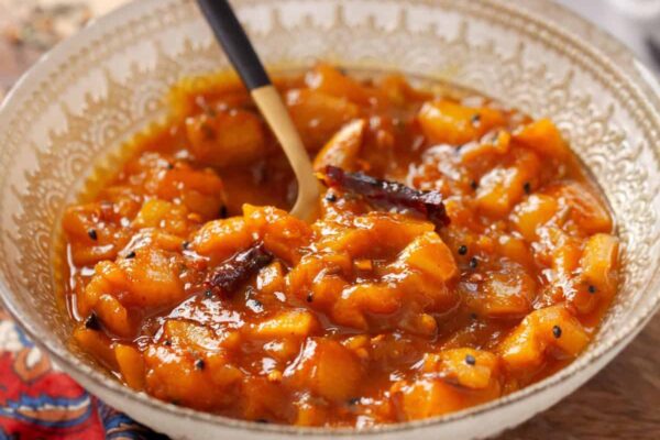 A bowl of mango chutney with a spoon, placed on a wooden board. The chutney is vibrant orange with visible chunks of mango and seeds.