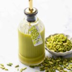 Glass bottle with green pistachio syrup and a cute tag tied on with twine, with a bowl of slivered pistachios on the side.
