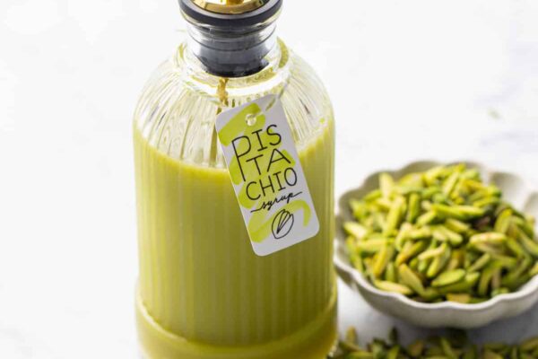 Glass bottle with green pistachio syrup and a cute tag tied on with twine, with a bowl of slivered pistachios on the side.
