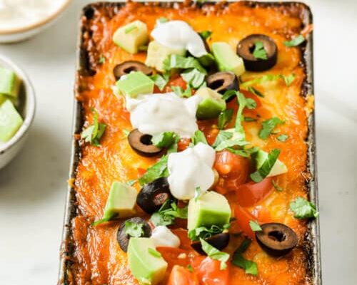 A loaf pan full of low carb lasagna, topped with golden brown baked cheese, olives, avocado, tomato, and sour cream.