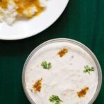 lauki ka raita in a glass bowl sprinkled with roasted cumin powder and coriander leaves on a dark green tray with a plate of dal and rice on top left side.