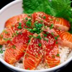 Close-up of a donburi bowl bursting with glossy salmon zuke, sprinkled with sesame and scallions, nestled against vibrant green shiso leaves.