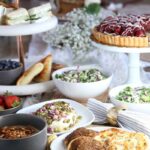 Side view of several white plating with bread, salads, cheese dome, tart and sanwiches