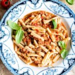 A blue and white vintage bowl with pasta in a fresh tomato sauce topped with basil. The bowl is on a travertine surface with cherry tomatoes and basil around.