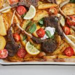 A tray of roasted lamb chops with golden potato wedges, cherry tomato halves, lemon slices, fresh mint leaves, and dollops of white sauce, all garnished and ready to serve.