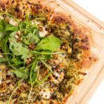 Pesto chicken pizza with goat cheese and pine nuts topped with arugula on a cutting board.