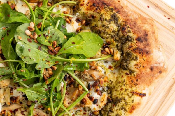 Pesto Chicken Pizza with Goat Cheese & Toasted Pine Nuts Pesto chicken pizza with goat cheese and pine nuts topped with arugula on a cutting board.