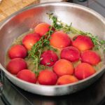 A stainless steel pan on the stovetop contains whole peaches and sprigs of fresh herbs simmering in the liquid.
