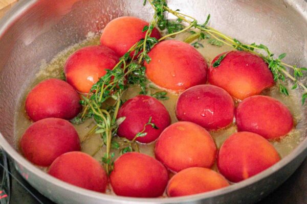 Roasted Apricots with Honey and Thyme A stainless steel pan on the stovetop contains whole peaches and sprigs of fresh herbs simmering in the liquid.