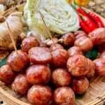 Sai krok Isan Thai sausages on a bamboo basket, with a backdrop of chilies, ginger, and cabbage.
