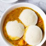 three idli on top of a layer of sambar in a white bowl with coconut chutney in a small bowl on top left.