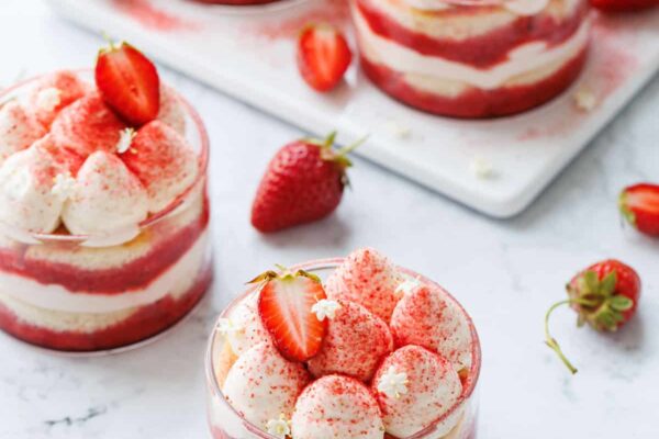 Strawberry Elderflower Tiramisu in individual clear ramekins, on a white marble background with two gold spoons and strawberries.