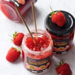 Three jars of homemade Strawberry Passionfruit Jam, one jar open with two gold spoons stuck inside, and a few fresh strawberries scattered around.